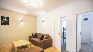 Un salon moderne avec un canapé marron, une table basse en bois et un petit pouf. Une porte donne accès à une autre pièce et un luminaire est fixé au mur.