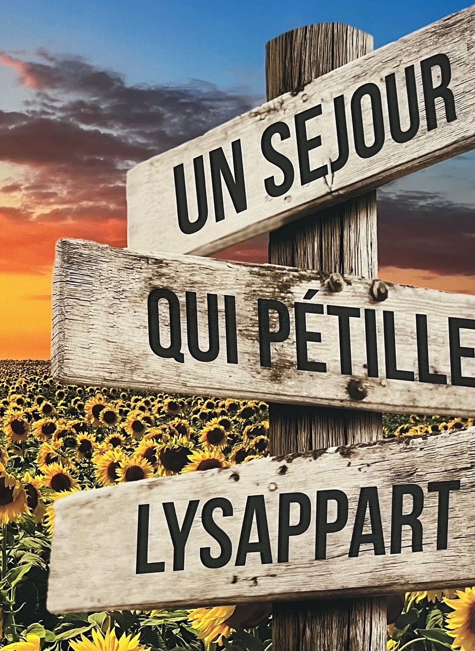 A wooden sign with the text "UN SEJOUR QUI PETIT LYSAPPART" against a sunset sky and sunflower field.