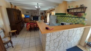 A kitchen with a stone counter, a dining area with a table and chairs, and a living room with a TV. - House in the countryside, with large expanses of woods and fields. (Goujounac)
