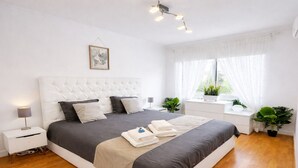 A modern bedroom with a tufted headboard, a large bed with grey bedding, a bedside table with a lamp, a dresser, and two potted plants. - Exclusive gallery apartment overlooking the golf course / beach (Charneca de Caparica)