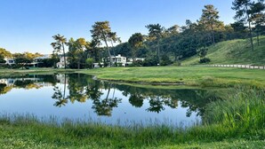 A serene landscape with a pond reflecting trees and buildings, surrounded by lush greenery and a clear sky. - Exclusive gallery apartment overlooking the golf course / beach (Charneca de Caparica)