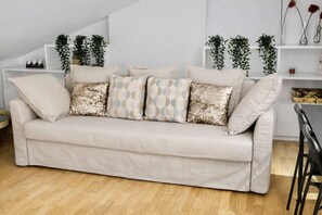 A neutral-toned sofa with patterned cushions, a wooden floor, and a white wall with shelves holding plants and decorative items.