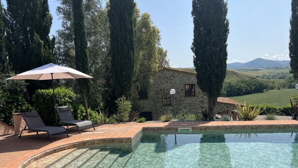 Una piscina limpida con gradini per accedervi, circondata da un patio in pietra. Si vedono due lettini e un ombrellone. Sullo sfondo, un edificio in pietra con una finestra e un lampione.
