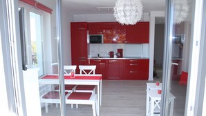 A modern kitchen with red cabinets and white countertops. There is a dining area with white furniture and a glass door leading to another room.