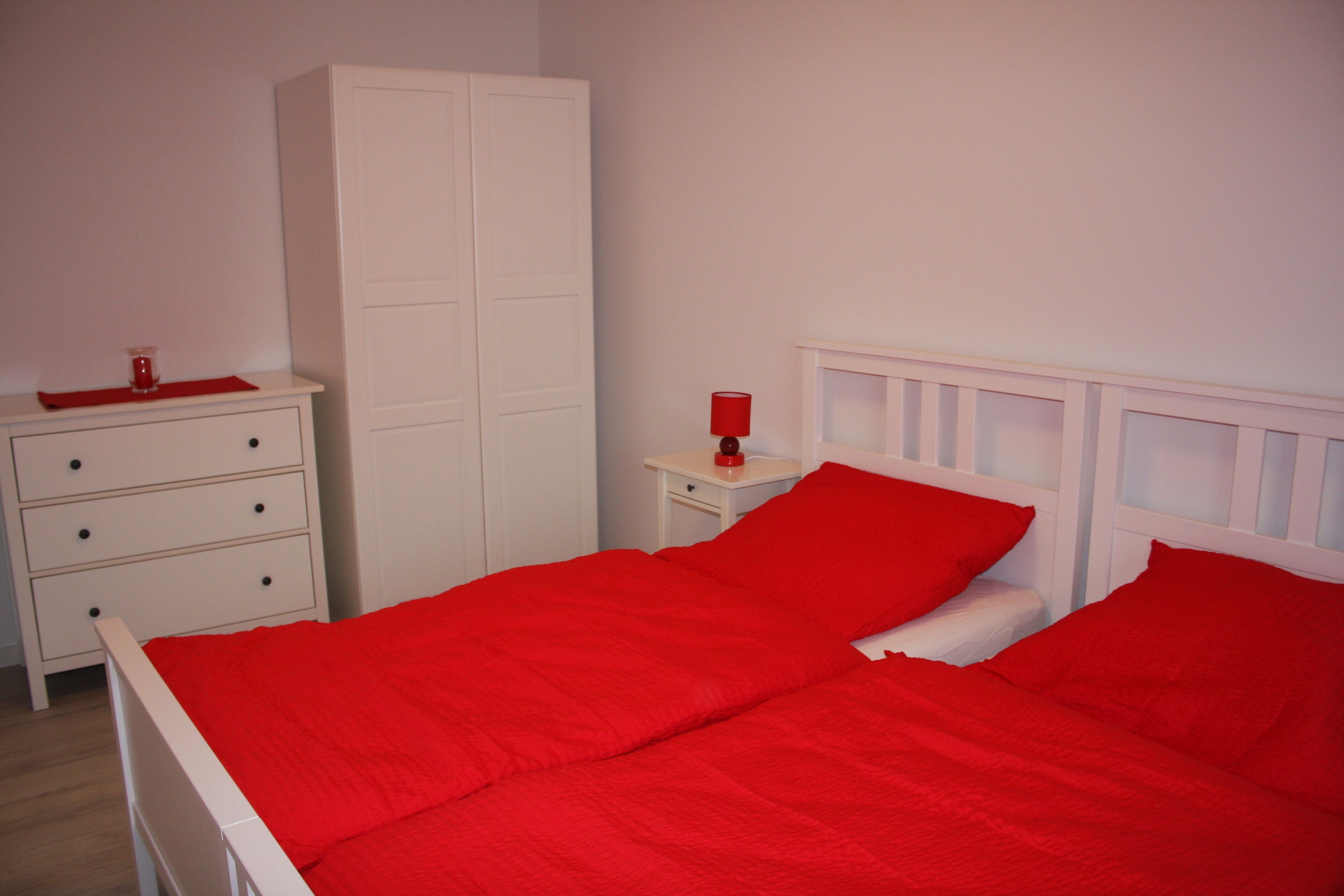 A bedroom with a white bed, red bedding, a white wardrobe, and a nightstand with a red lamp.