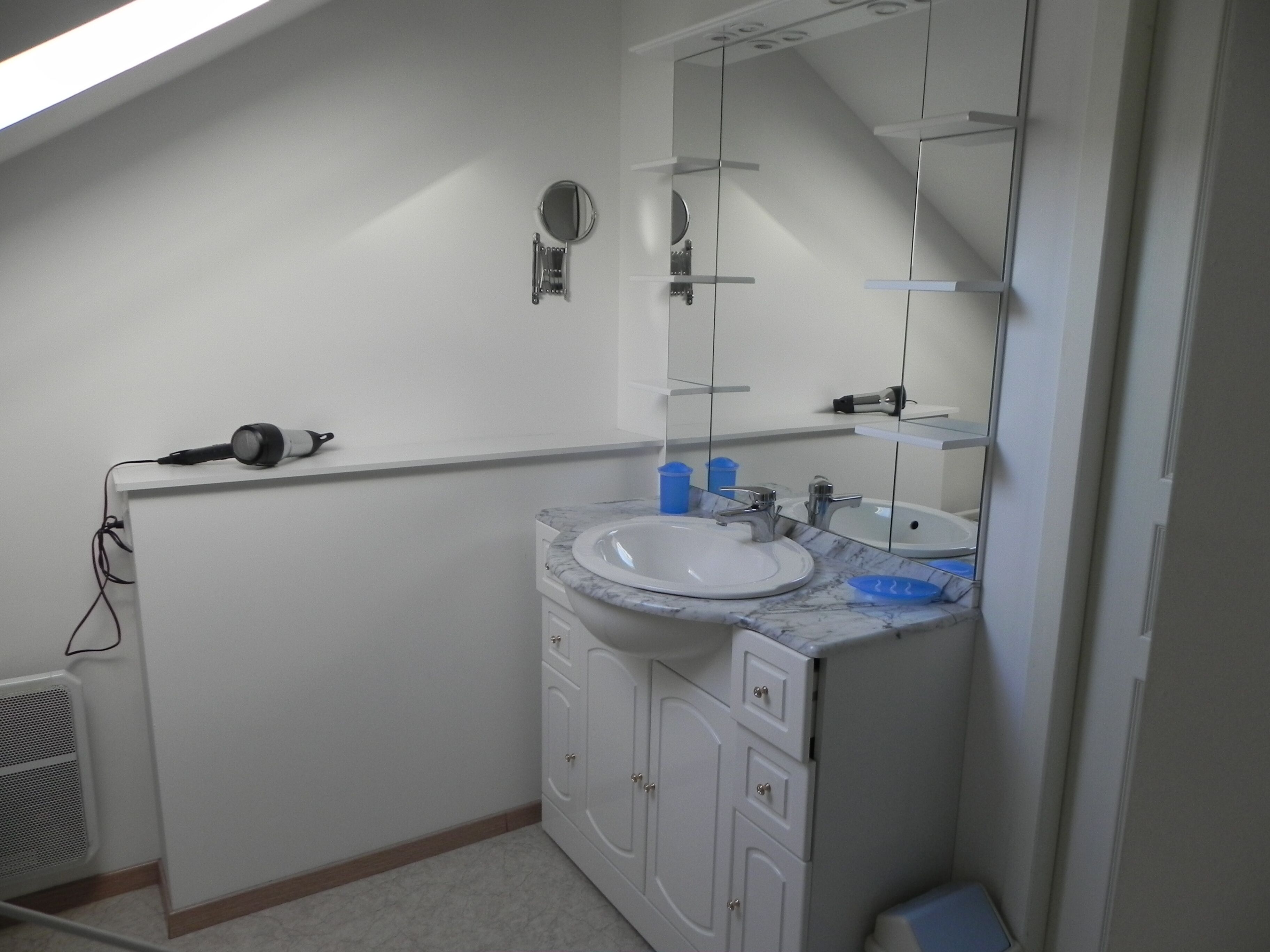 Une salle de bain avec un lavabo blanc, un miroir et des armoires. On y trouve un sèche-cheveux sur le comptoir et une grille d’aération sur le mur.