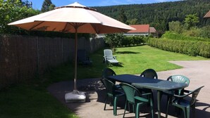 An outdoor patio with a table and chairs, a large umbrella, and a view of a forested hillside.