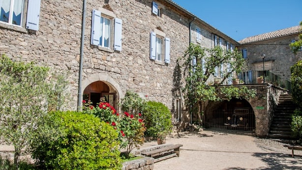 Un bâtiment en pierre, doté d’arcades au niveau des portes et de fenêtres avec volets, entouré de verdure et bordé d’une cour pavée.