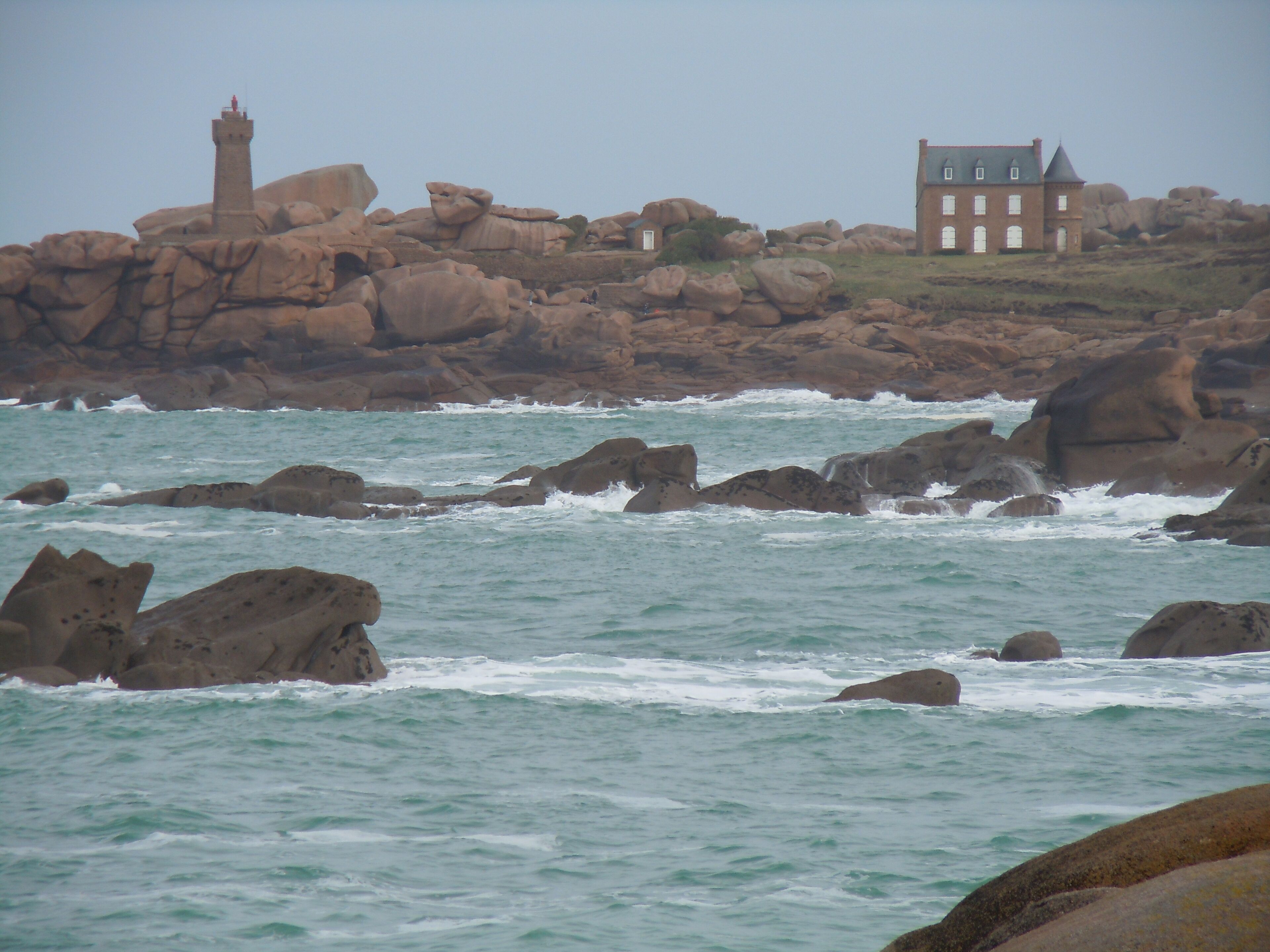 A coastal landscape with large boulders, a lighthouse, and a building with a steep roof.
