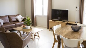 Een moderne woonkamer met een bruine leren bank, een houten salontafel, een televisie en een eethoek met een houten tafel en stoelen.