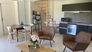 Een moderne woonkamer met een houten eettafel, twee bruine fauteuils, een salontafel met een vaas, een boekenkast en een keuken met een magnetron en oven.