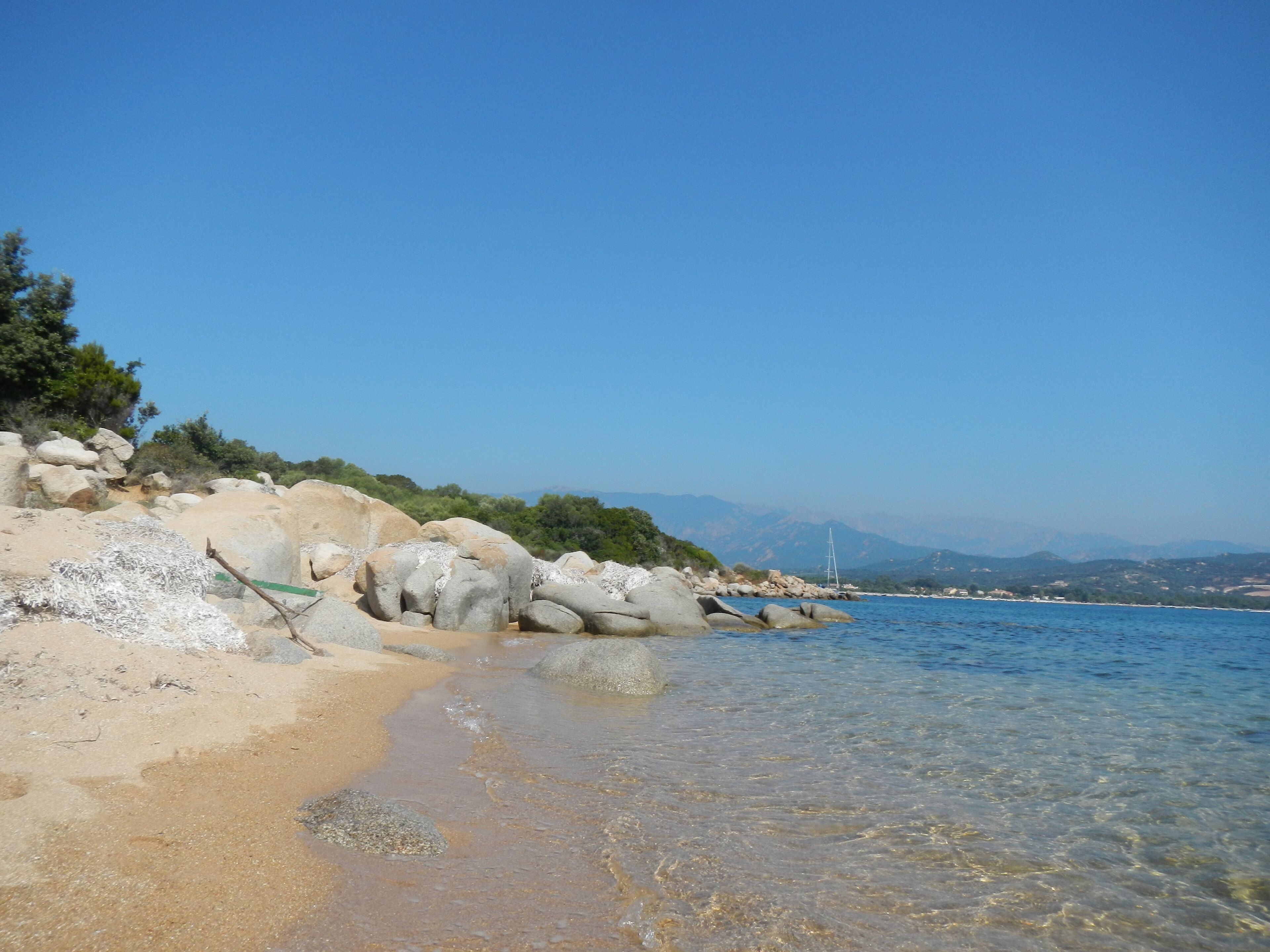 A beach with clear shallow waters, large rocks, and distant mountains.