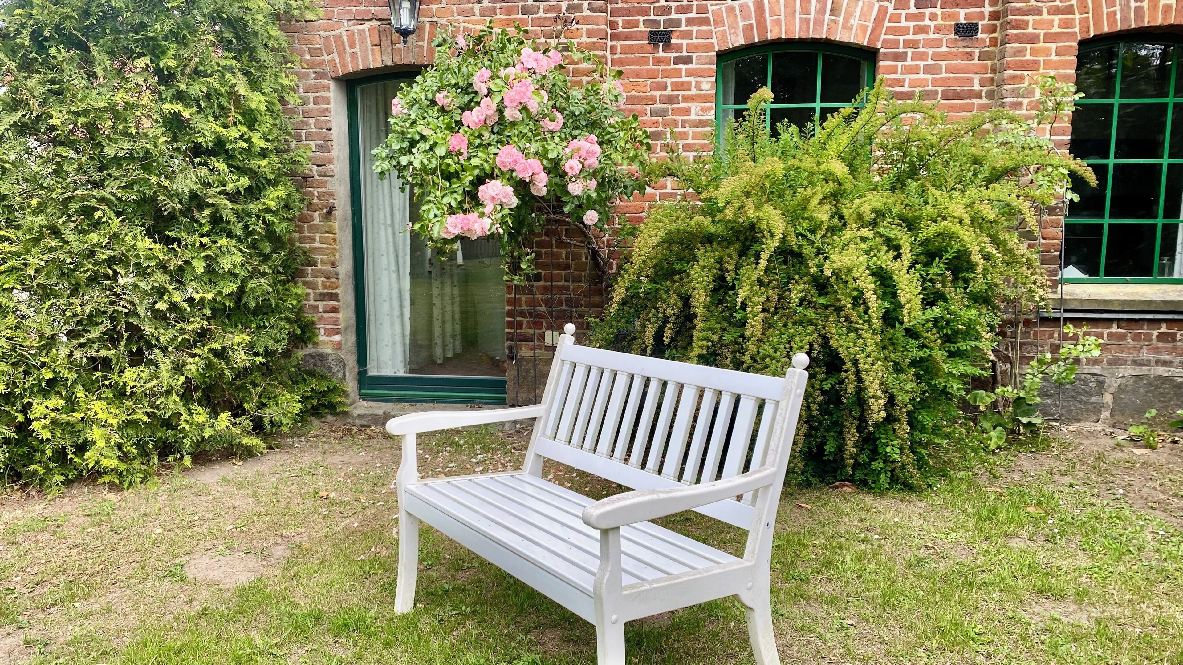 Un banc blanc dans un jardin, avec un bâtiment en briques en arrière-plan.