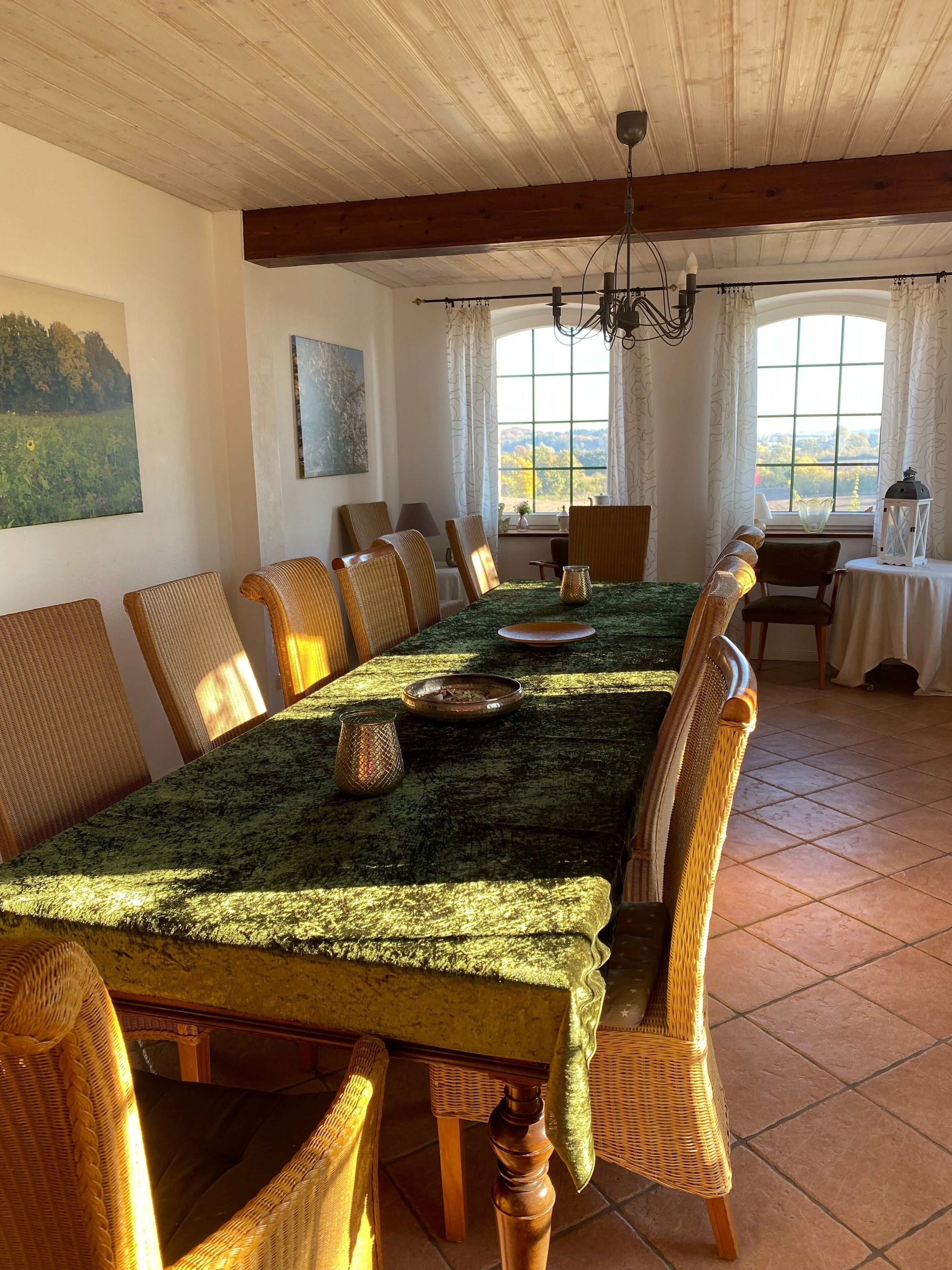 A dining area with a long table, wicker chairs, a chandelier, and large windows with curtains.