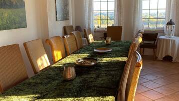 A dining area with a long table, wicker chairs, a chandelier, and large windows with curtains.