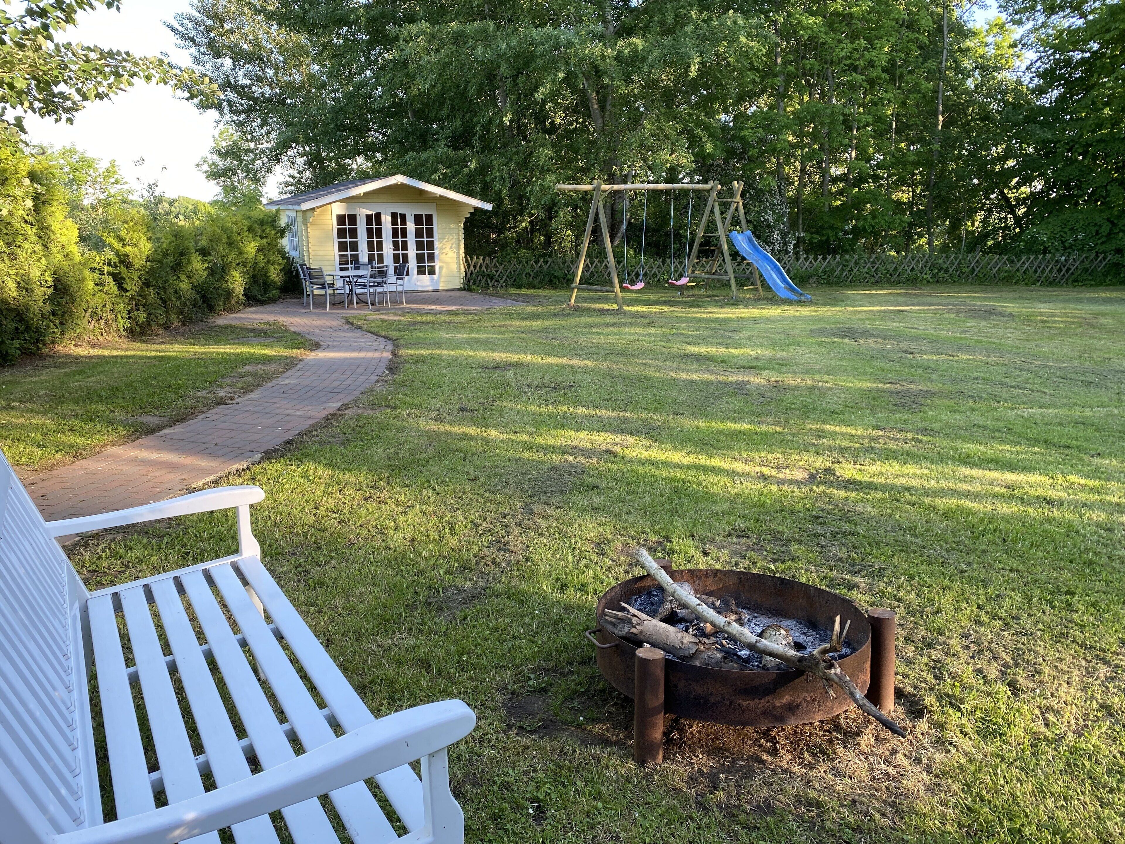 A backyard with a white bench, a small wooden house, a swing set, and a fire pit.