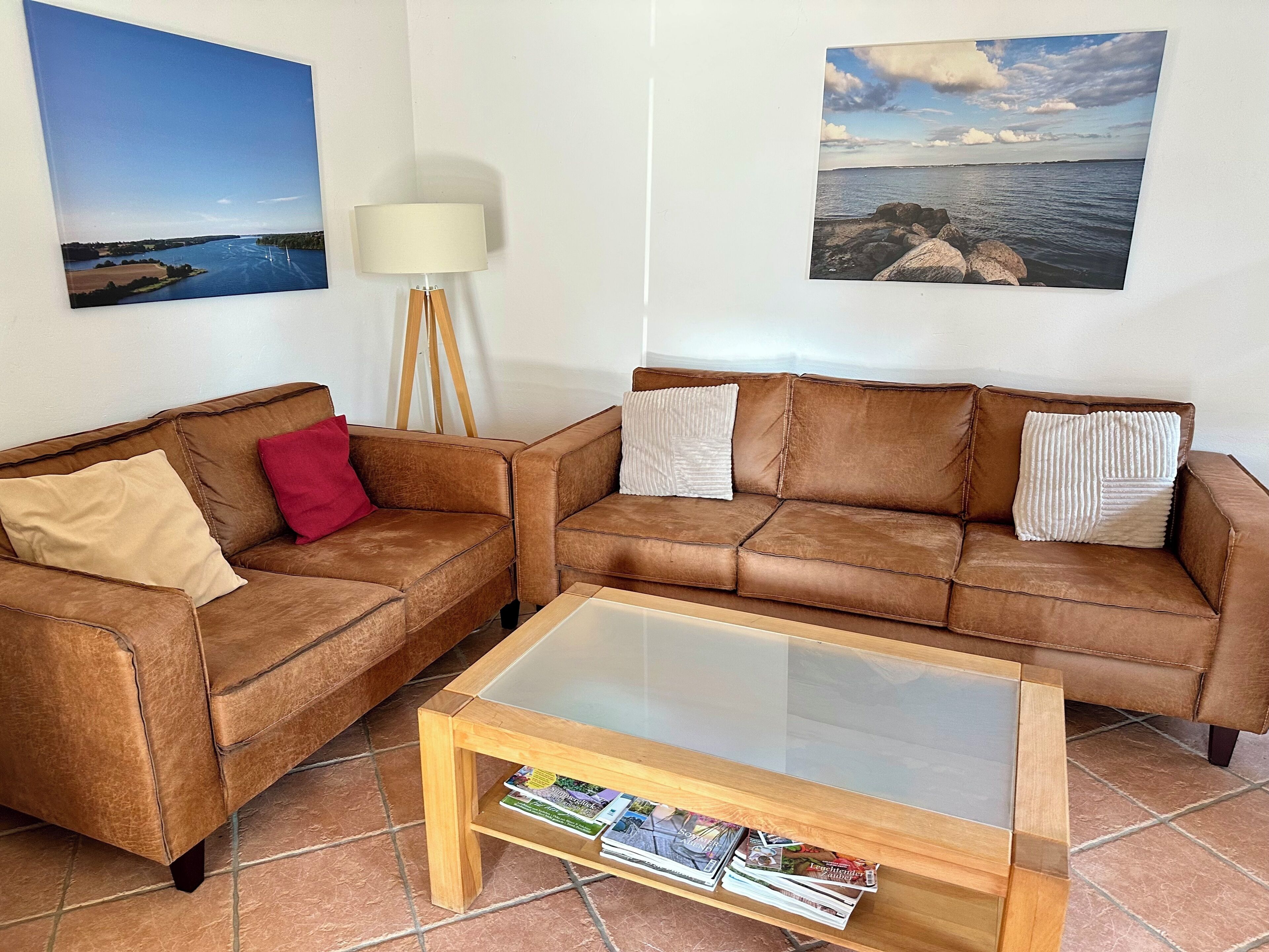 A living room with two brown leather sofas, a glass-top coffee table with magazines, and two wall-mounted pictures depicting landscapes.