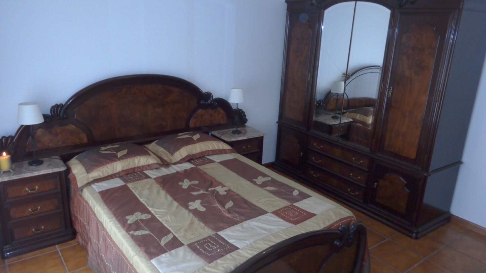 A bedroom with a wooden bed, a nightstand with a lamp, a dresser with a mirror, and a tiled floor.