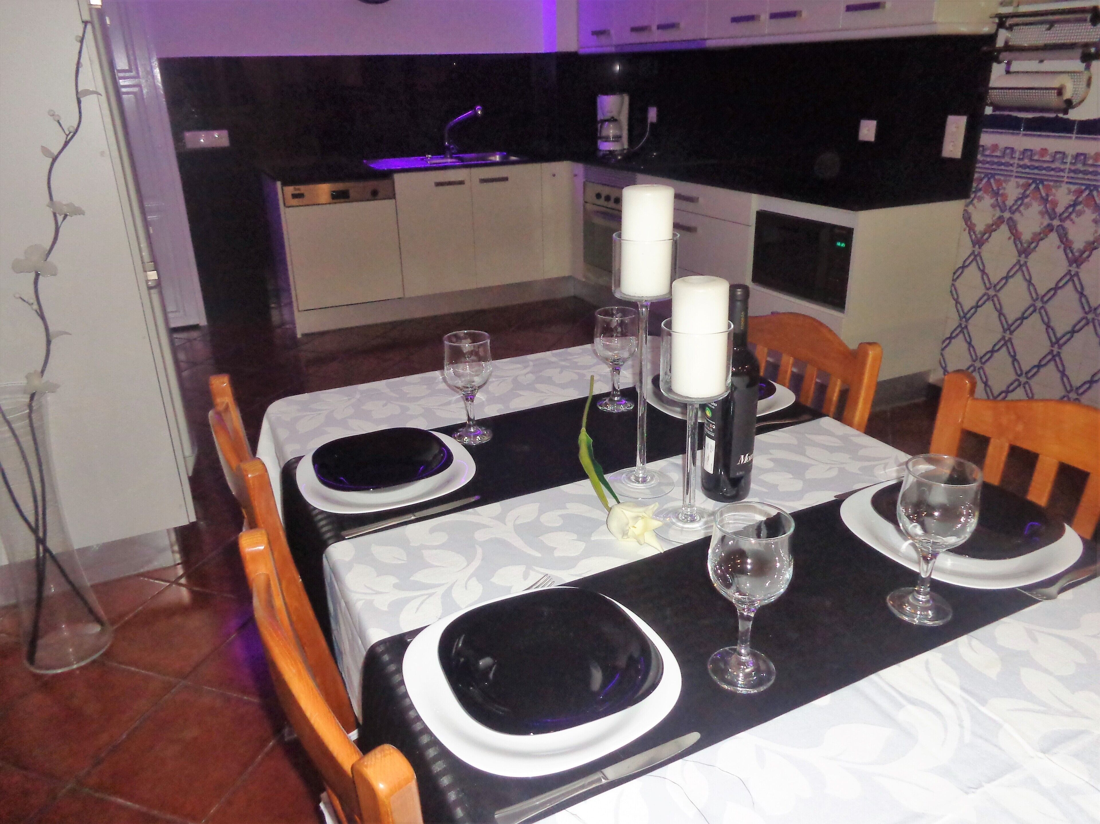 A dining table set with plates, glasses, and candles, with a kitchen in the background.