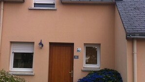 Ein zweistöckiges Gebäude mit einer beigen Fassade, einer Holztür und einem kleinen Fenster mit Fensterläden. Vor der Tür befindet sich ein blau blühender Busch.