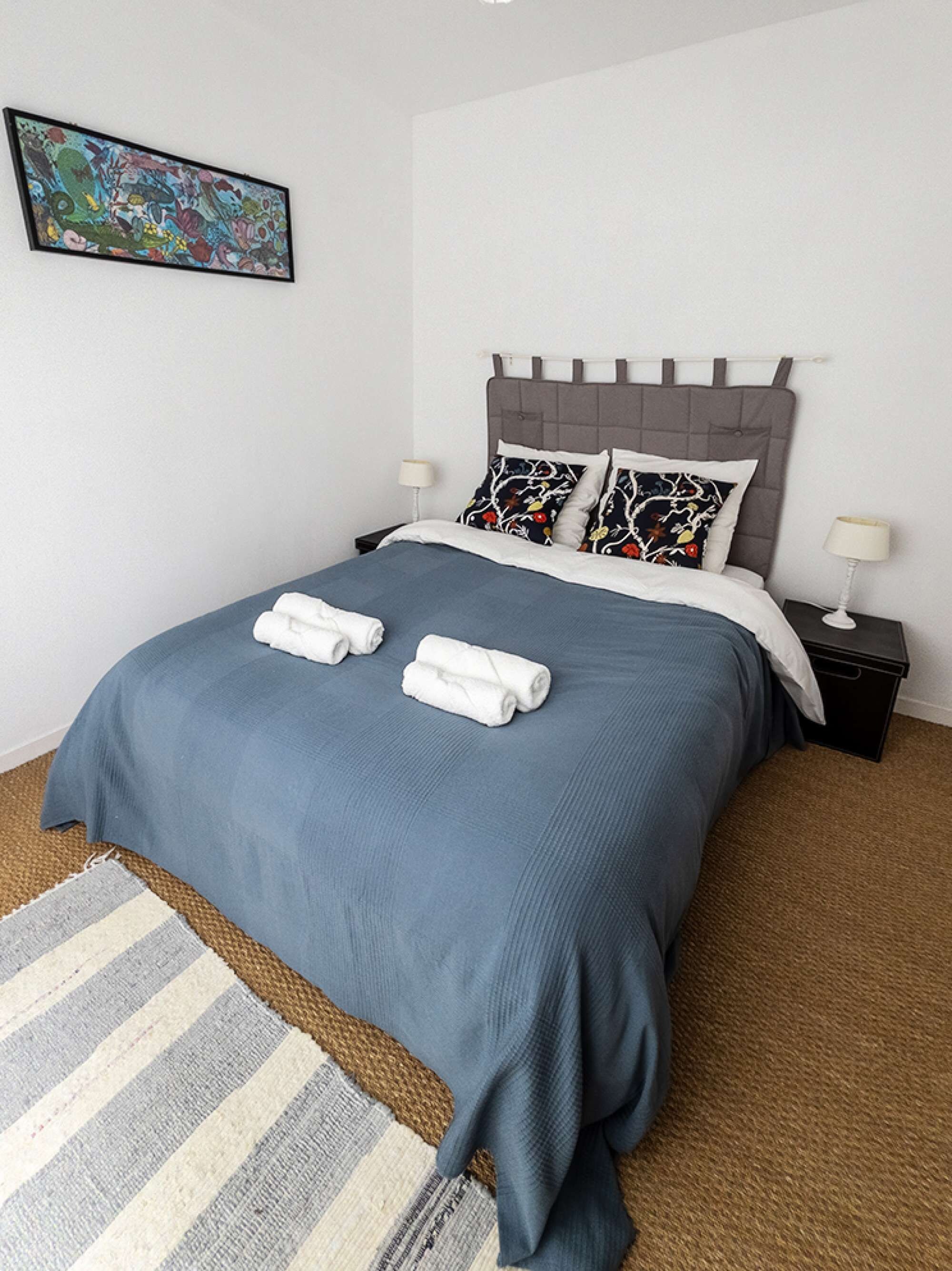 A neatly made bed with a grey headboard, two pillows with a floral pattern, and two rolled towels.
