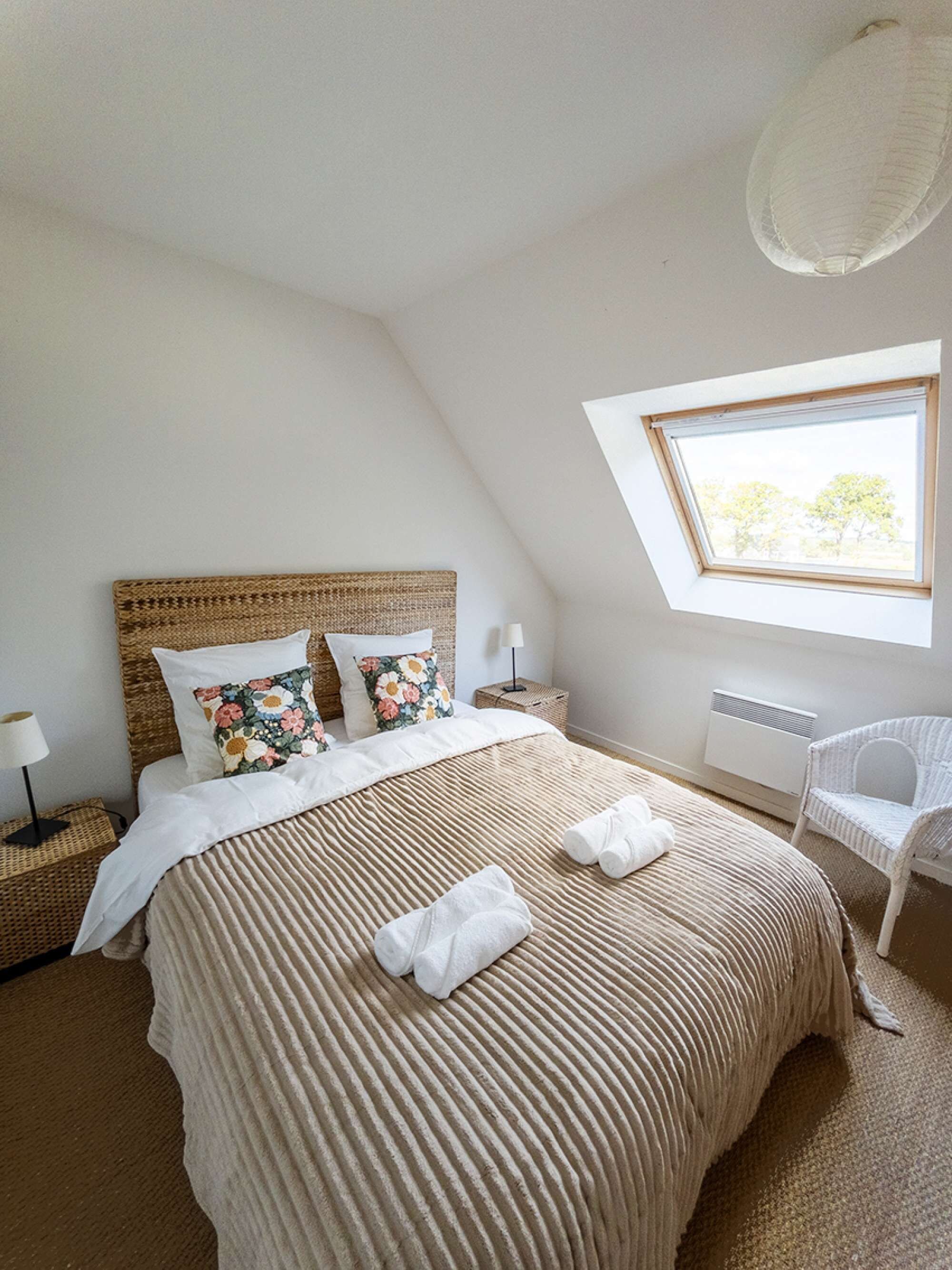 Ein Schlafzimmer mit schrägem Dach, einem großen Bett mit gestreifter Bettdecke, zwei Kissen, einem Nachttisch mit Lampe, einem Sessel und einem Fenster mit Blick auf Bäume.