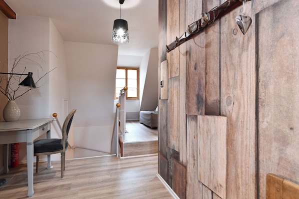 A room with a wooden door, a dining table with chairs, a hanging light fixture, and a view of another room with a bed.