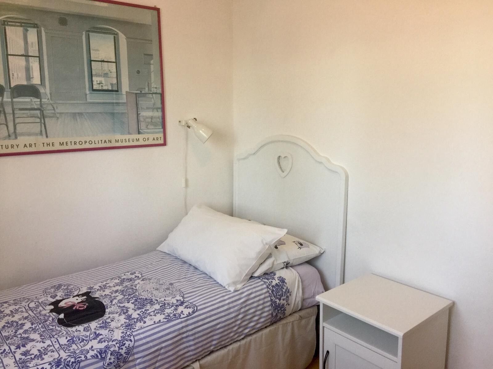 A small, neatly made bed with a heart-shaped headboard, a bedside table, and a framed picture of a museum on the wall.