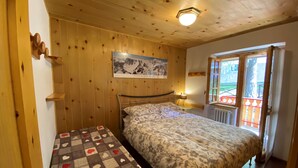 A bedroom with a wooden ceiling, a bed with floral bedding, a bench with a heart-patterned blanket, and a mountain painting on the wall.