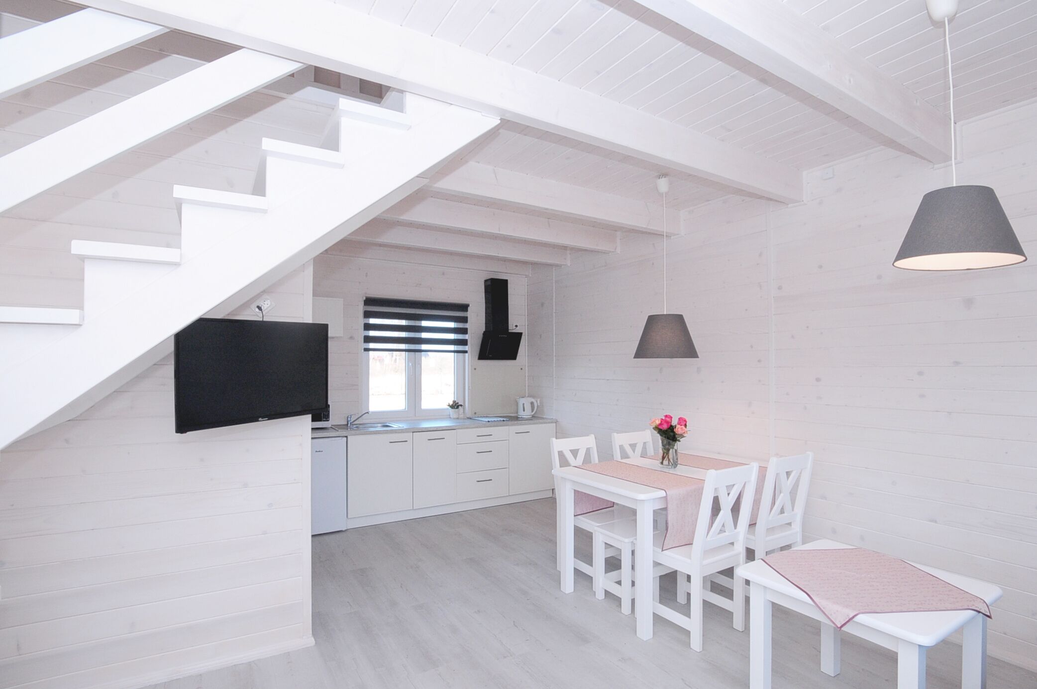 A modern kitchen with white cabinetry, a black TV mounted on the wall, and a dining area with white chairs and a table.