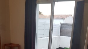 Un dormitorio con cama, una silla de madera, una mesita y un ventanal corredero que da a un balcón con cerca blanca y césped verde.