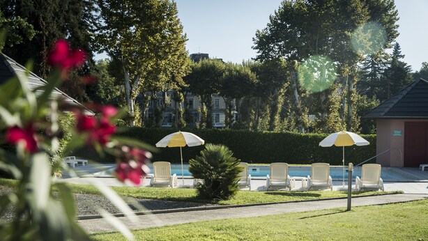 Un espace aménagé au bord de la piscine, avec des chaises longues et des parasols, entouré de verdure et d’arbres.