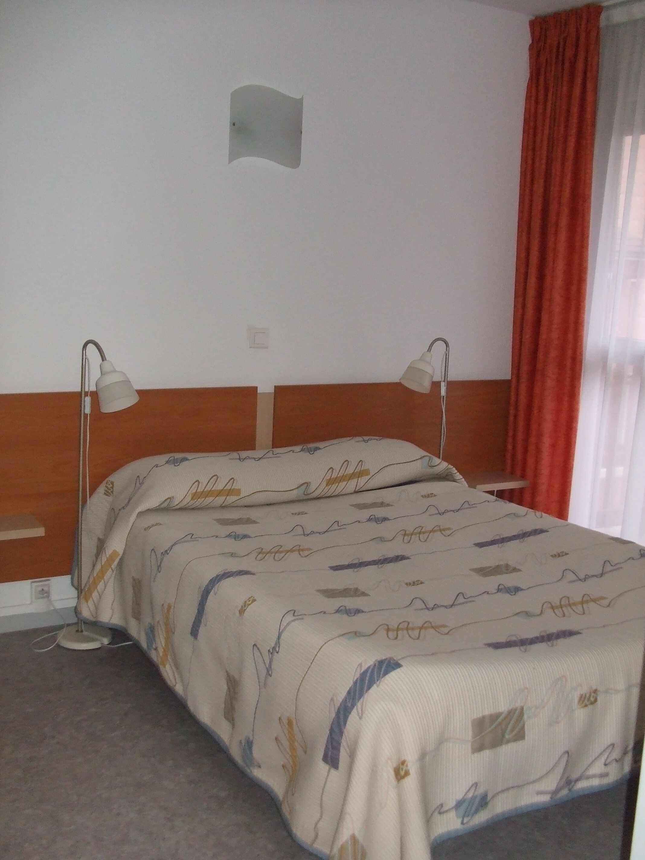 A hotel room with a bed, two bedside lamps, a red curtain, and a plain wall with a light switch.