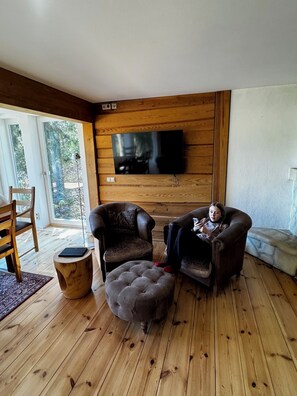 Ein gemütliches Wohnzimmer mit Holzboden, einem großen Flachbildfernseher an einer Holzwand, zwei Ledersesseln, einem kleinen Hocker und einem Holztischchen.