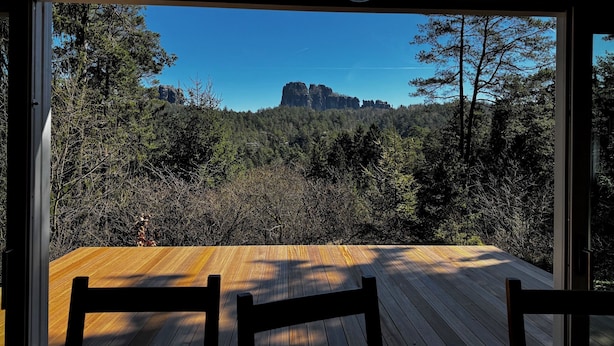 A wooden deck with chairs, a view of a forest and rock formation through large windows.