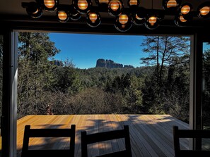 A wooden deck with chairs, a view of a forest and rock formation through large windows.
