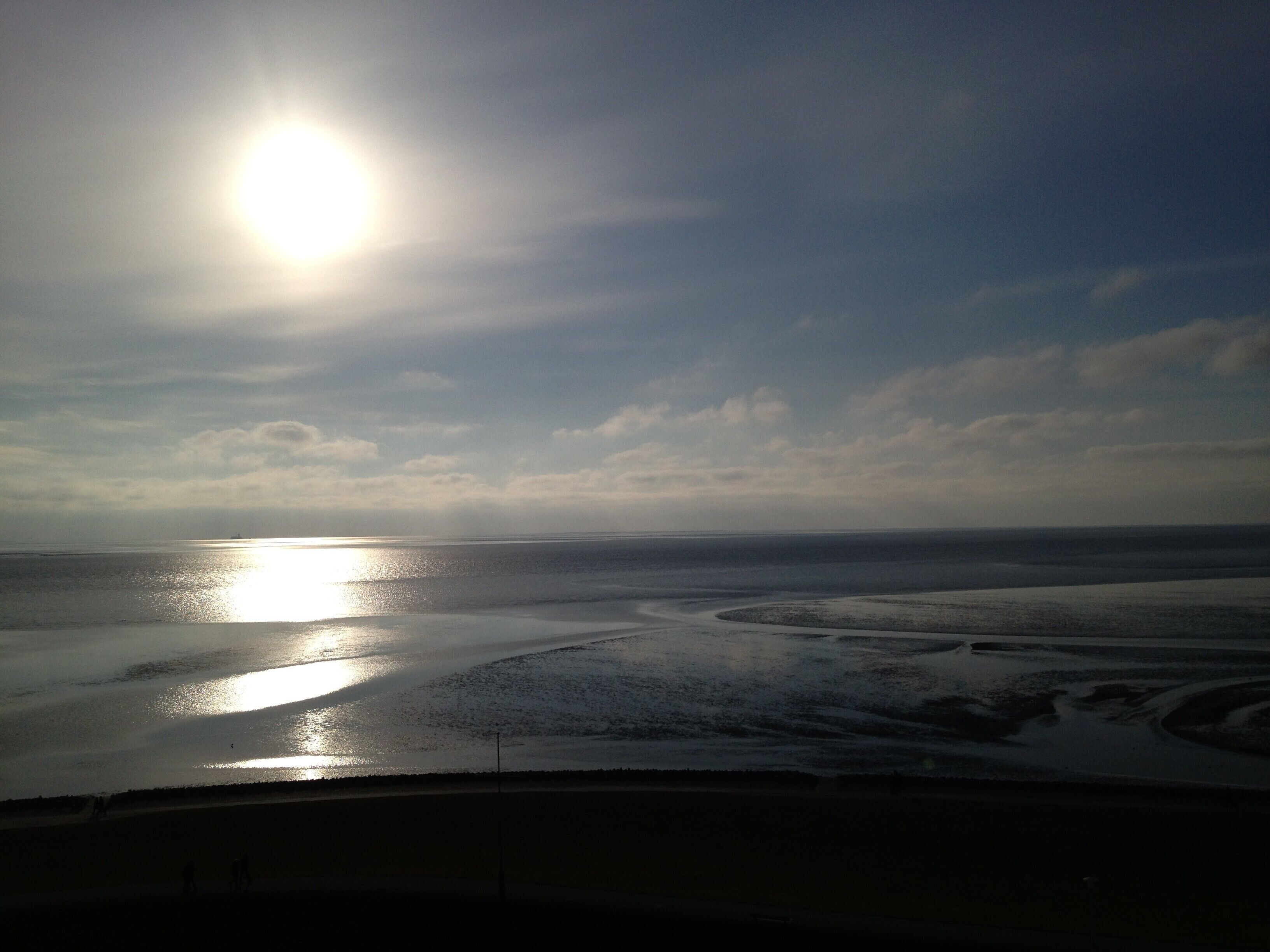 A beach with the sun reflecting on the water, a clear sky, and a distant view of the ocean.