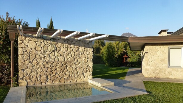 Un muro de piedra con una pérgola, una piscina y un césped bien cuidado.