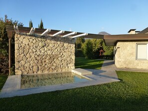 Een stenen muur met een pergola, een zwembad en een verzorgd gazon.