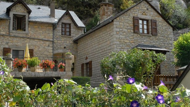 A stone building with a balcony, surrounded by greenery and flowers. - GITE IN THE HEART OF THE GORGES DU TARN (La Malène)
