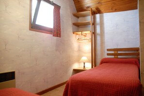 A bedroom with two beds, a window with a curtain, a bedside table with a lamp, and a wardrobe with hanging clothes.
