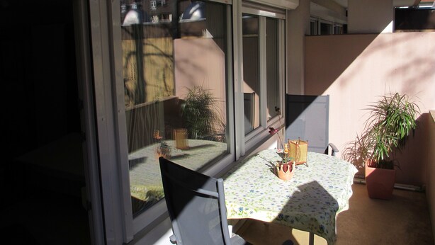 A balcony with a table and chairs, potted plants, and a view of a residential area through large sliding glass doors.