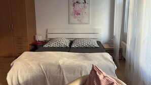 A bedroom with a bed, a nightstand, a lamp, a window with curtains, and a wooden wardrobe.