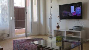 A modern living room with a glass coffee table, a flat-screen TV mounted on the wall, a potted plant, and a bookshelf.