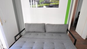 A grey sofa bed with a black frame, a small potted plant, and two green slippers on a black glass coffee table.