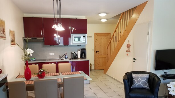 Een moderne keuken met rode kastjes, een magnetron en een koffiezetapparaat. Er staat een eettafel met stoelen en een bloevas. In de woonkamer is een zwarte leren fauteuil en een televisie te zien.