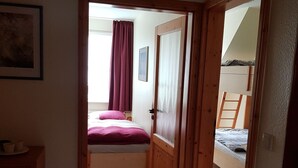 Een smalle gang die leidt naar een slaapkamer met een bed, een raam met gordijnen en een deur.
