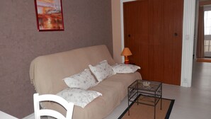 A living room with a beige sofa, patterned pillows, a glass coffee table, a white chair, and a wooden door. - Apartment in very quiet building (Royan)