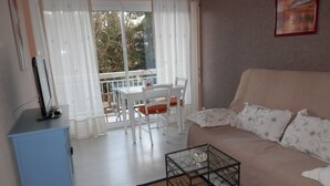 A living room with a sofa, a glass coffee table, a TV on a stand, a dining table with chairs, and a view through the window. - Apartment in very quiet building (Royan)