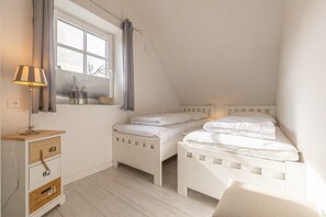 Ett sovrum med två vita sängar, en byrå i trä med lådor, en lampa och ett fönster med gardiner.